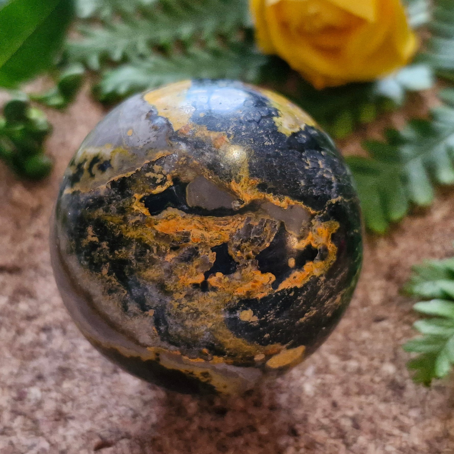 A striking polished stone sphere featuring dark tones with vivid golden-yellow and rusty orange patterns, resembling volcanic or mineral-rich textures. It rests on a cork surface with green ferns and a yellow flower in the softly blurred background, creating a natural and earthy atmosphere.
