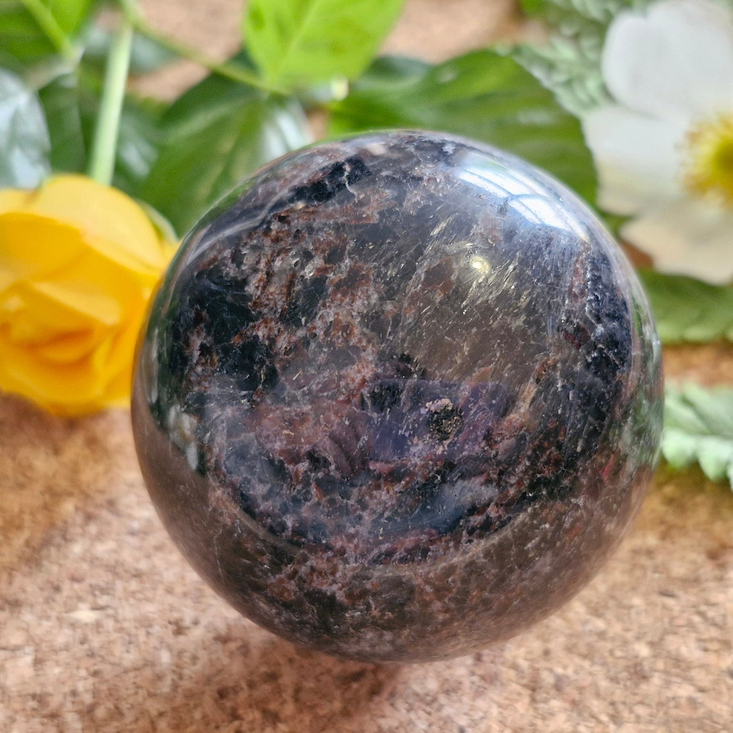 Polished dark sphere with rich reddish-black patterns, resting on a cork surface surrounded by yellow and white flowers and green leaves.
