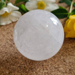 Polished clear quartz sphere resting on a cork surface, surrounded by white and yellow flowers and green leaves. The crystal has soft internal textures and subtle rainbows.