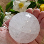 Hand holding a clear quartz sphere, showing its smooth surface and internal clarity. A soft floral background adds a calming, natural feel.