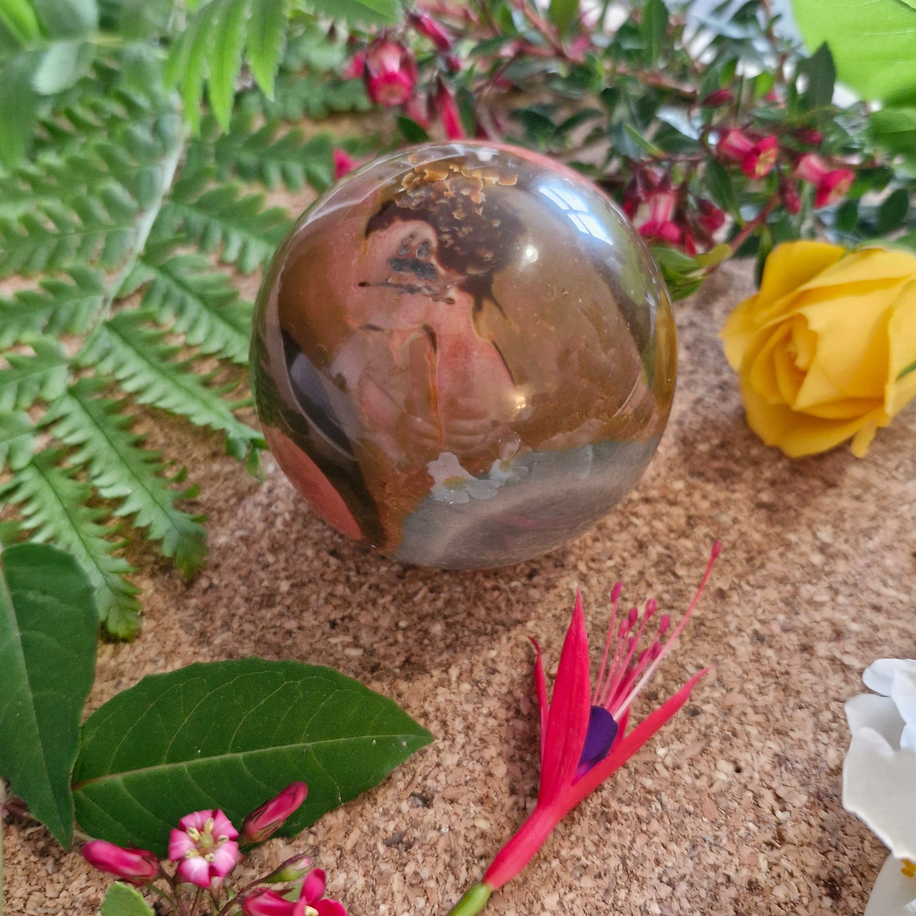 A beautifully polished orb with earthy tones of brown, pink, and grey, resting on a cork surface. It's surrounded by vibrant greenery, a yellow rose, and small red and pink flowers, creating a colourful, natural setting.
